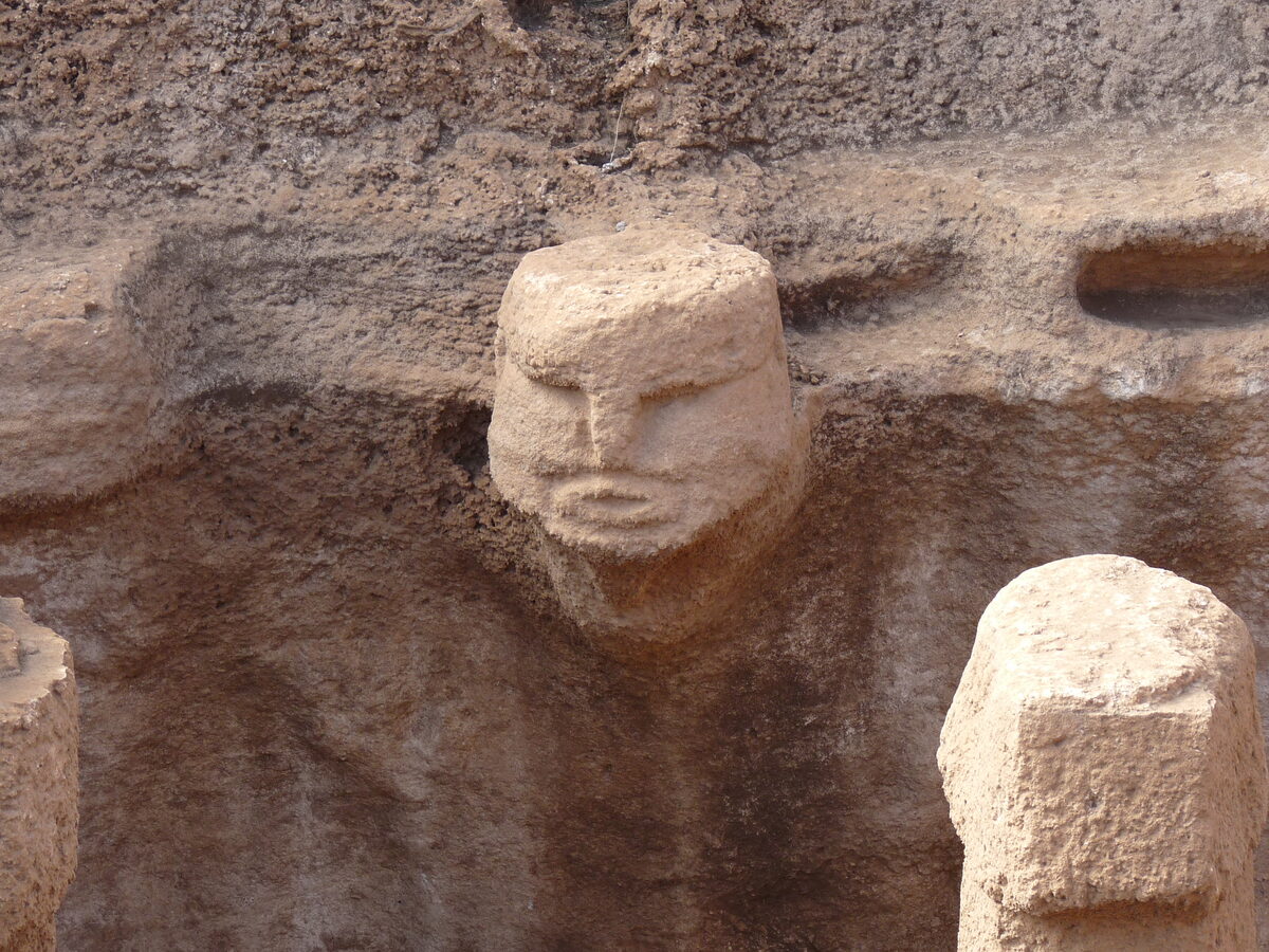 Tête humaine sculptée en haut-relief émergeant de la paroi rocheuse dans la salle aux piliers de Karahan Tepe, le cou allongé et le regard tourné vers la pièce.